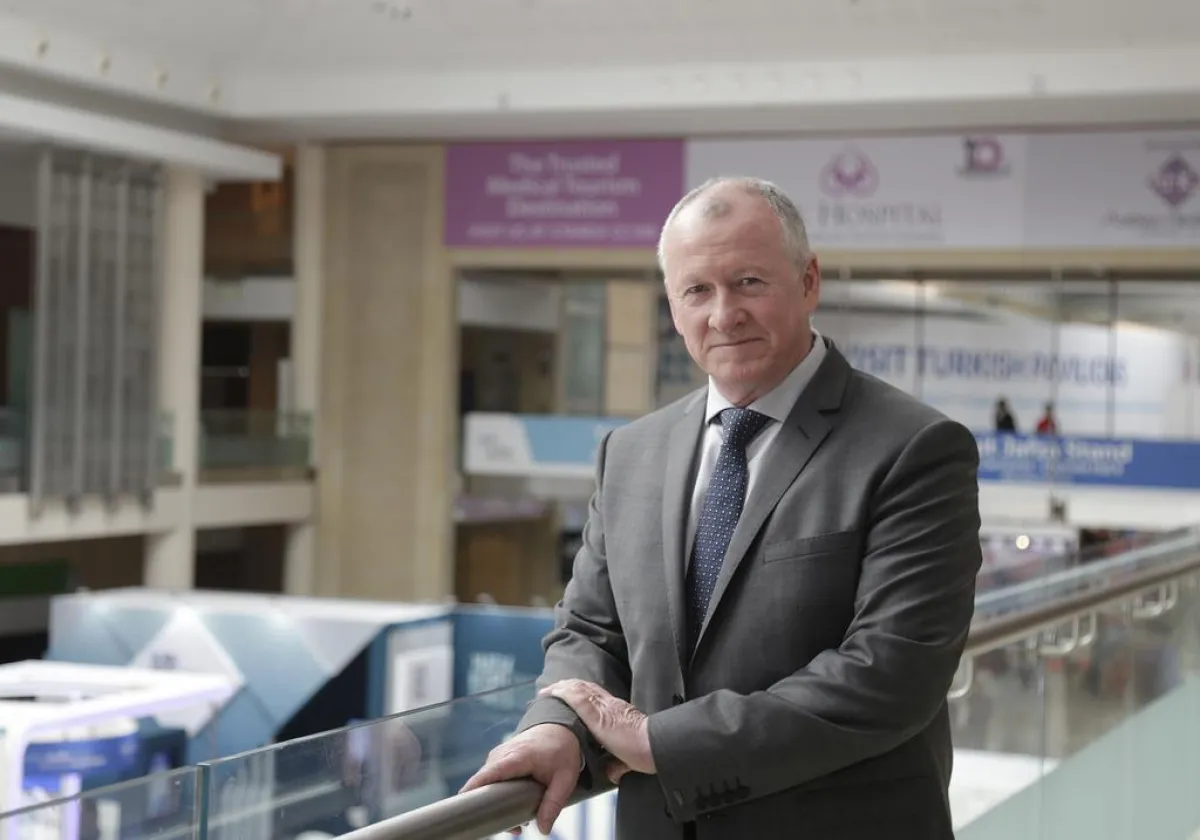 A middle-aged man in a gray suit and tie stands leaning on a railing inside a modern building with banners hanging in the background.