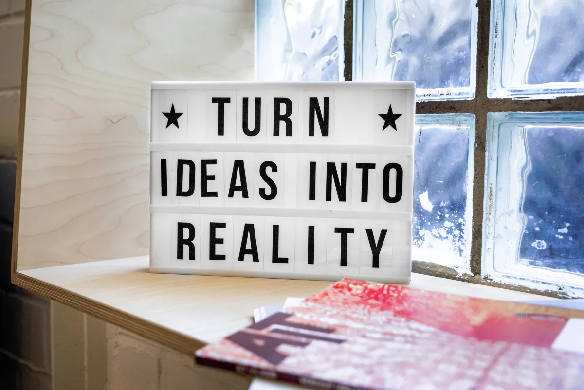 A lightbox sign reading "TURN IDEAS INTO REALITY" placed on a shelf by a window with translucent glass.