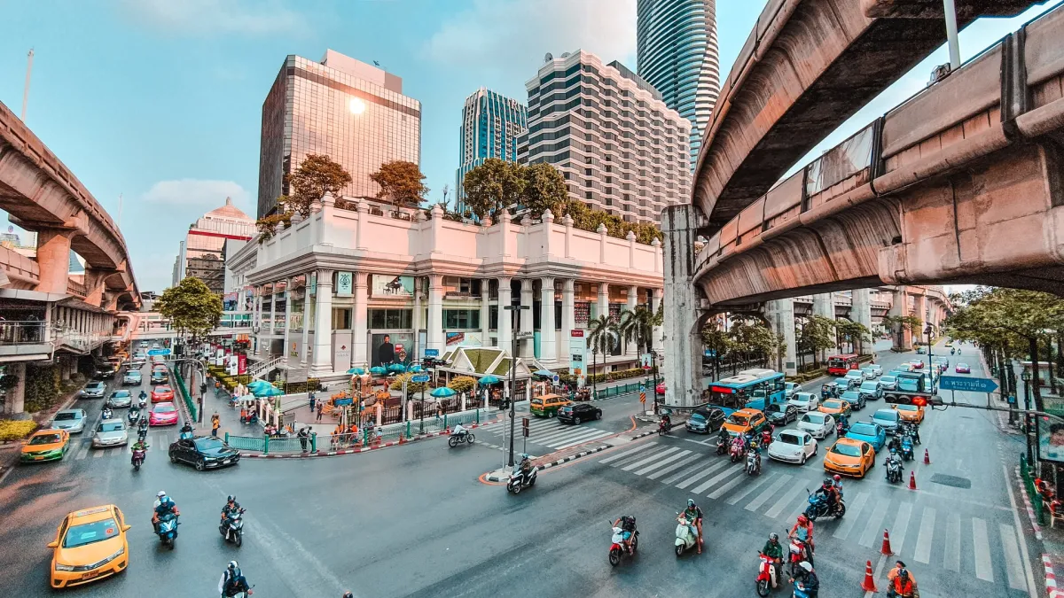 A bustling city intersection featuring a mix of cars and motorcycles, with elevated train tracks and modern skyscrapers in the background.