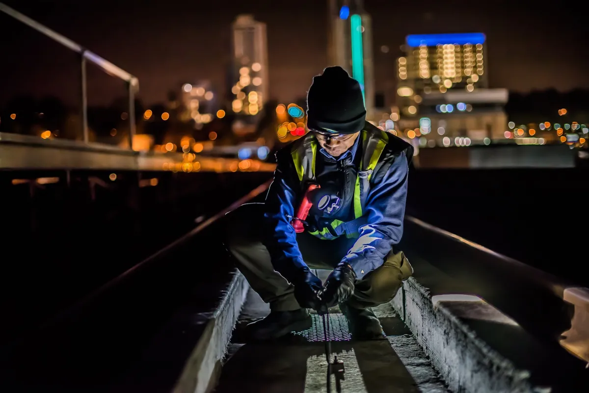 A worker in high-visibility gear inspects equipment on an illuminated rooftop at night with a cityscape in the background.