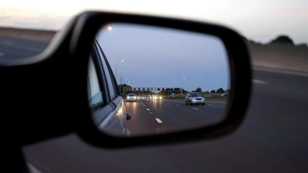 View of a highway reflected in a car's side mirror during evening, showing vehicles illuminated by their headlights.