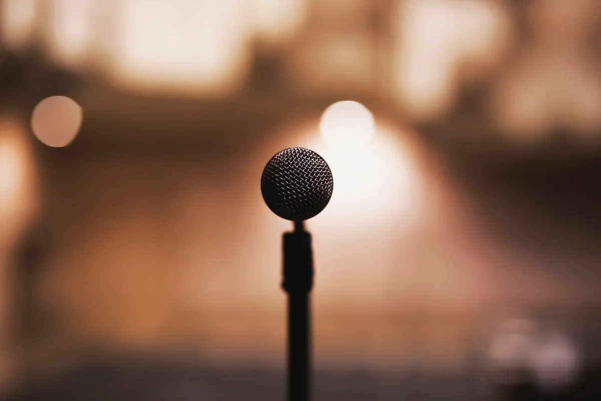 A microphone stands in focus against a softly blurred background with warm, glowing lights.