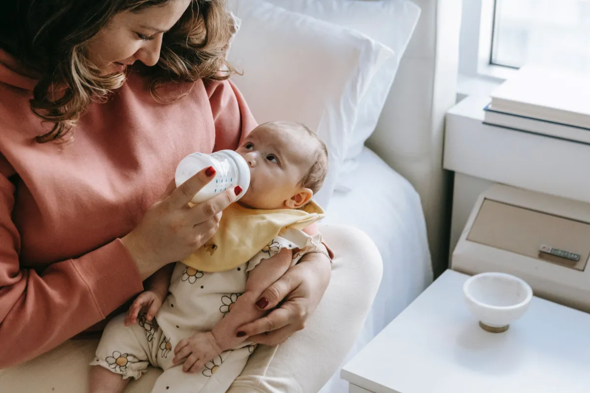 A woman in a pink sweatshirt feeding a baby with a bottle while sitting near a window.