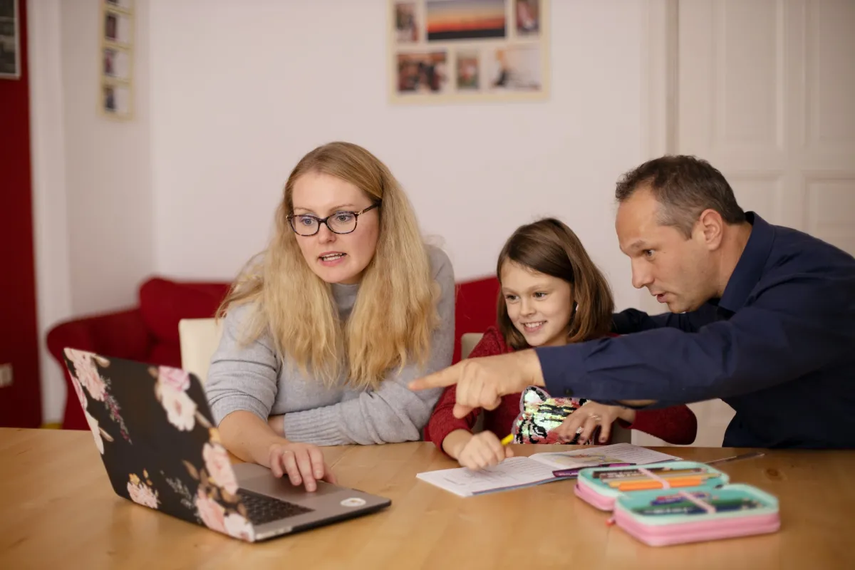 A woman, a man, and a young girl sit at a table with a laptop, engaging in a family activity together.