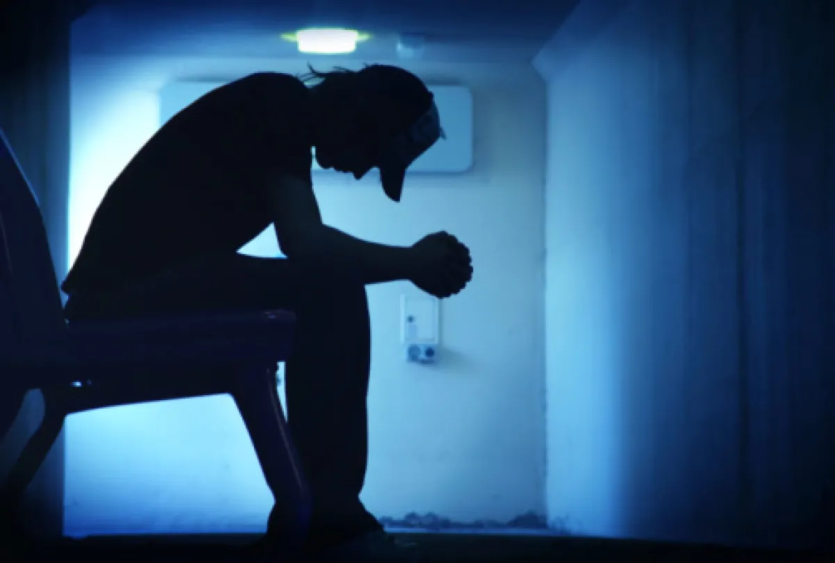 A silhouette of a person sitting with their head bowed down in a dimly lit, blue corridor.