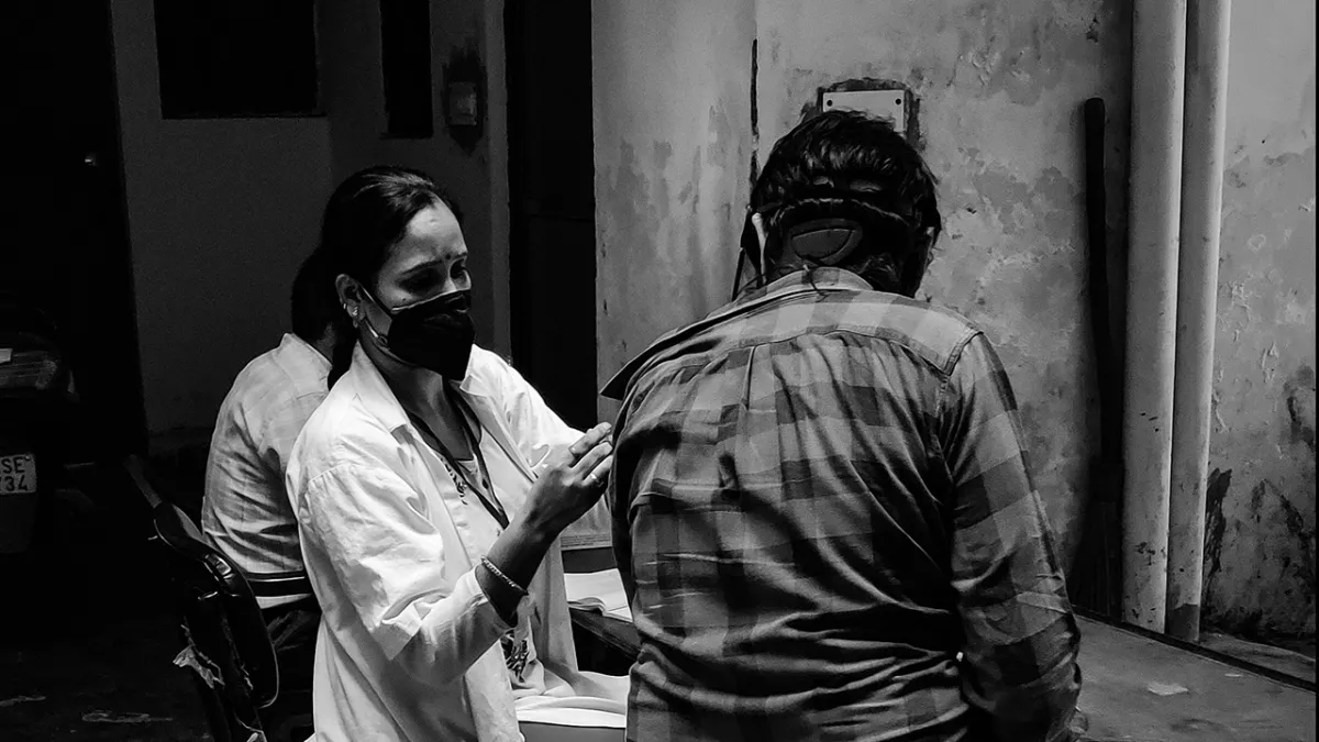 A healthcare worker in a white coat administers a vaccine to a man in a plaid shirt, both wearing masks, in a dimly lit setting.