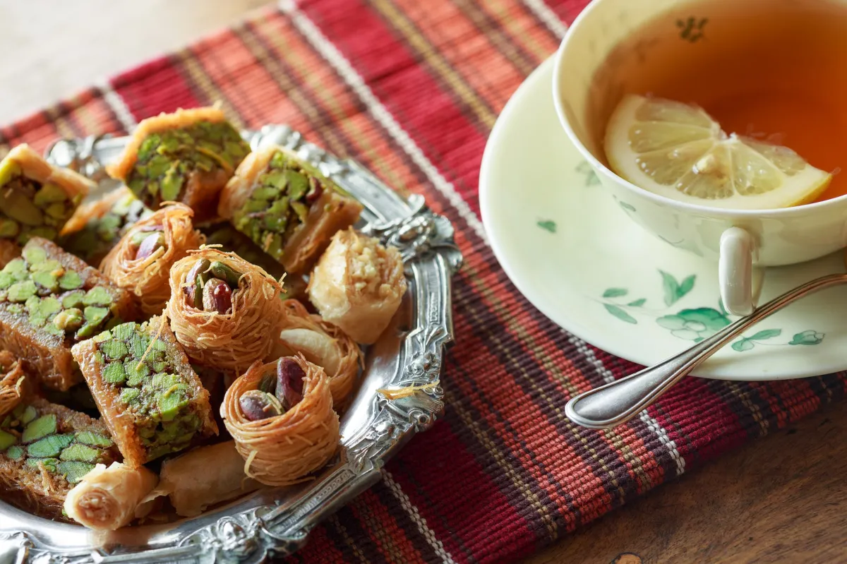 A variety of Middle Eastern pastries on a silver tray beside a cup of tea with lemon slice on a patterned tablecloth.