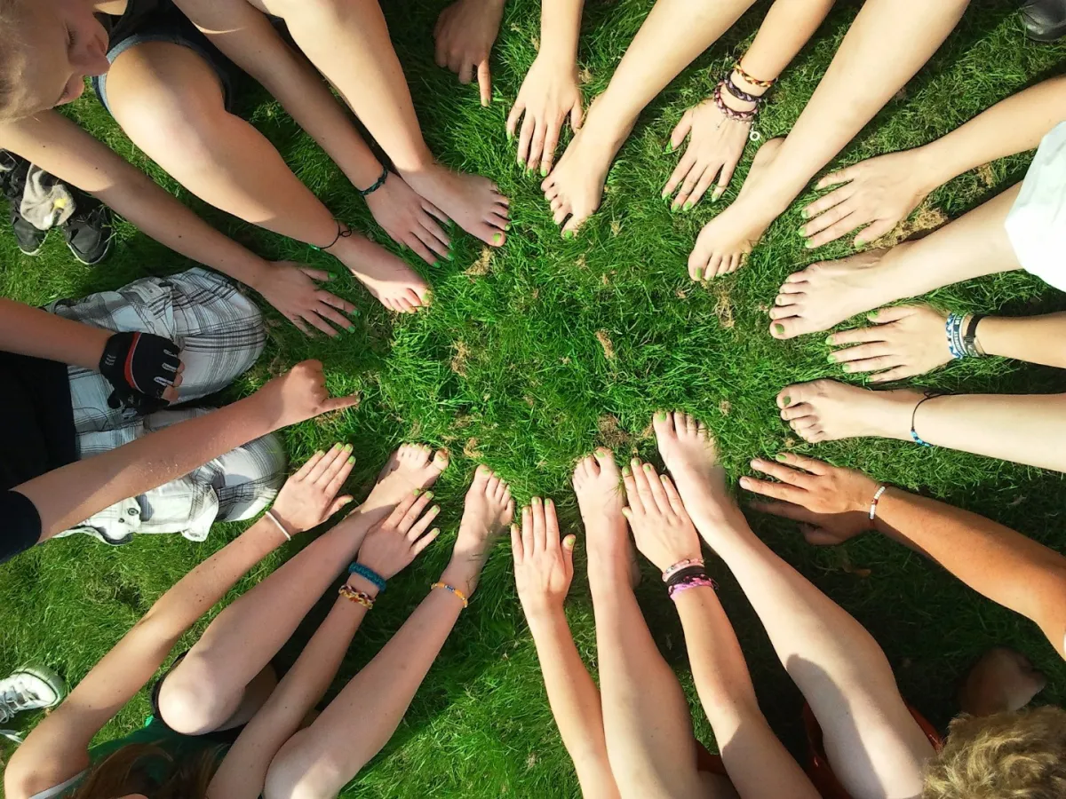 A group of people sitting in a circle on grass showing their bare feet and hands meeting in the center.