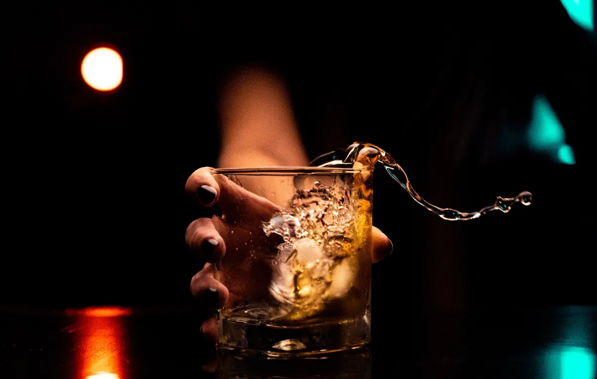 A hand tipping a glass with amber liquid, causing a splash against a dark, softly lit background.