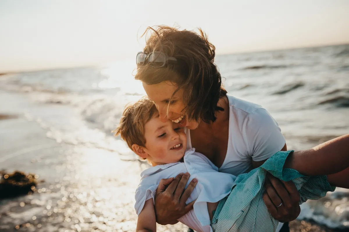 A woman tenderly embracing a young boy as they both smile near the ocean waves at sunset.
