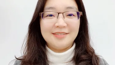 Portrait of a smiling woman with glasses, wearing a white turtleneck and a gray cardigan over a green top.