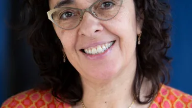 A smiling middle-aged woman wearing glasses, a vibrant orange and pink patterned jacket, and a gold necklace.