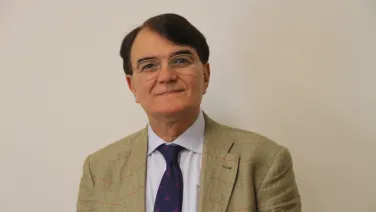 A middle-aged man wearing glasses, a beige checkered suit, and a blue tie with pink motifs, smiling at the camera against a plain beige background.