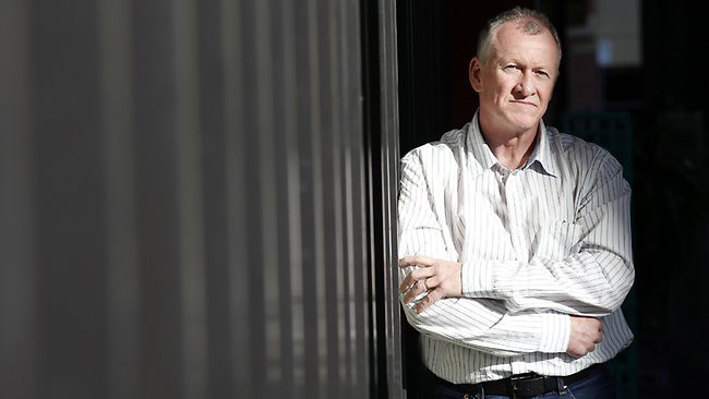 A man standing with crossed arms leaning against a doorway with vertical blinds, looking thoughtful.